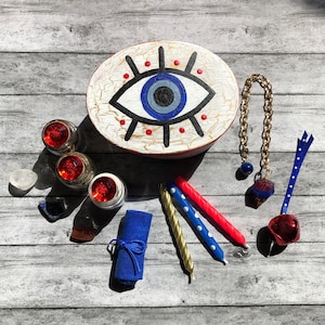May include: A collection of spiritual items including an oval box with an eye design, candles, crystals, and a gold chain with a charm. The box has a white background and a red border. Candles are red, blue, and gold. Crystals are visible.