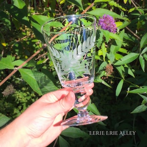 Op de afbeelding: Een helder glazen wijnglas met een gegraveerd ontwerp van bladeren en stelen. Het glas heeft een lange, slanke steel en een brede, ronde kom.