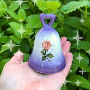 May include: A ceramic bell with a gradient finish transitioning from a deep purple base to a white center. The bell features a painted orange rose with green leaves and a heart-shaped handle at the top, designed for decorative display.