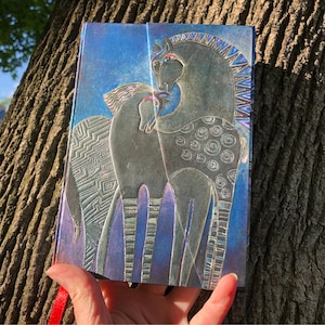 May include: A blue journal with an embossed design of two horses. The horses are gray with intricate patterns and details. The journal has a red ribbon bookmark and a textured cover. The background is a gradient of blue.