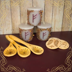 May include: Three white candles with a rune symbol and dried herbs around the edges are stacked. Two wooden spoons with flower designs and three wooden coasters with etched designs are in the foreground.
