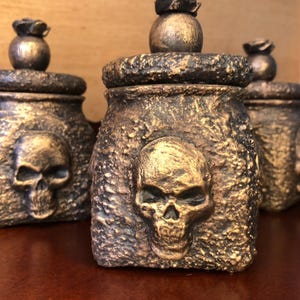May include: Three bronze-coloured, textured jars with skull designs. Each jar has a lid topped with a decorative sphere and a rose-like embellishment. The jars are arranged on a dark wooden surface.