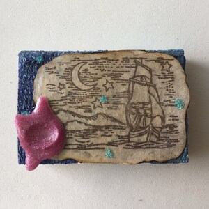 May include: A blue and brown decorative box with a vintage nautical scene of a ship sailing under a crescent moon and stars. The box has a pink star-shaped embellishment on the front.