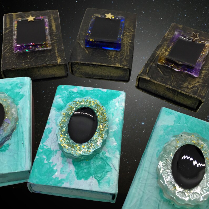 May include: An assortment of decorative boxes with unique designs. Some boxes are dark with glittery accents and star-shaped embellishments. Others are teal with oval-shaped, glitter-framed black stones. The boxes are arranged against a starry background.