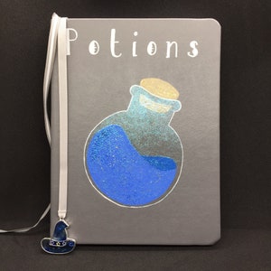 May include: Gray journal with a white ribbon bookmark and a blue potion bottle with a cork stopper. The journal cover has the word "Potions" written in white. A blue witch hat charm is attached to the ribbon.