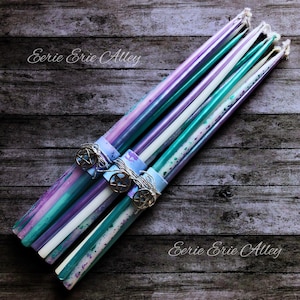 Pode incluir: Um conjunto de velas longas e finas em tons de roxo, azul-turquesa e branco com pingentes de pentagrama prateados. As velas são amarradas com barbante e têm um efeito salpicado, marmorizado. O texto "Eerie Erie Alley" é visível duas vezes na imagem.