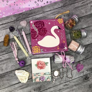 May include: An assortment of spiritual and decorative items, including a pink swan painting, candles, crystals, small jars of herbs and salts, a seashell, a small mirror, and a heart-shaped pendant. The items are arranged on a gray wooden surface.