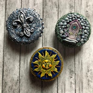 Könnte beinhalten: Drei dekorative, runde Objekte mit einzigartigen Designs. Eines zeigt eine silberne Fleur-de-Lis auf einer strukturierten blauen Oberfläche. Ein anderes zeigt eine Feentür. Das dritte hat ein lächelndes Sonnengesicht auf blauem Hintergrund.