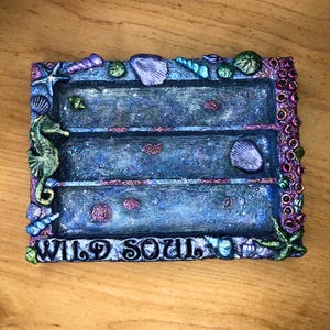 Puede incluir: Una bandeja rectangular de tres niveles con temática marina. La bandeja es principalmente azul con detalles de purpurina y presenta adornos de conchas marinas e hipocampos. Las palabras "WILD SOUL" están en la parte inferior.