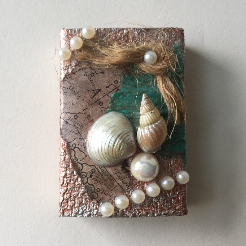 May include: A small square canvas with a textured, metallic gold background. The canvas is decorated with seashells, pearls, and a piece of brown twine. The seashells are white and brown, and the pearls are white. The canvas has a vintage map design in the background.