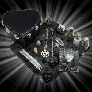 May include: A black heart-shaped box with silver studs, a silver chain with a pentagram pendant, a white crystal, a silver bell, three small glass bottles with silver lids, two seashells, two black candles, a black heart-shaped stone, and a black and silver heart-shaped tray with a silver heart on top, all on a black and silver fabric with a star pattern.