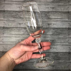 Pode incluir: Uma taça de champanhe transparente com uma haste decorativa e uma base redonda. A haste apresenta um design semelhante ao cristal. O copo é segurado em ângulo contra um fundo de madeira cinza.