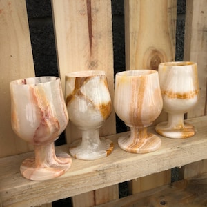 May include: Four onyx wine glasses with a unique, natural pattern of white, brown, and orange hues. The glasses have a rounded bowl shape and are set on a short stem and a circular base. The glasses are displayed on a wooden surface.