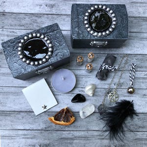 May include: An assortment of mystical items arranged on a wooden surface. Two gray decorative boxes with silver accents are featured, along with a candle, crystals, a feather, and a pentagram necklace. Small jars and a scroll are also included.