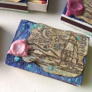 May include: A small blue box with a decorative paper insert featuring a ship sailing on a moonlit sea. The paper is decorated with glitter and stars. A pink, glittery embellishment is on the left side of the box.