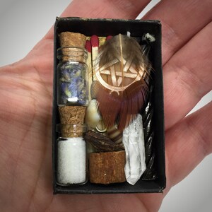 May include: A black matchbox filled with small glass bottles, dried flowers, matches, feathers, a crystal, and other natural elements. The box is held in a hand, showcasing the arrangement of items.
