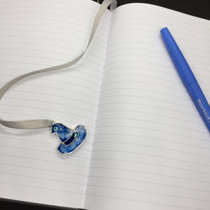 May include: A lined notebook with a gray ribbon bookmark and a blue witch hat charm. A blue pen with the text "Paper Mate" is on the right side of the notebook.