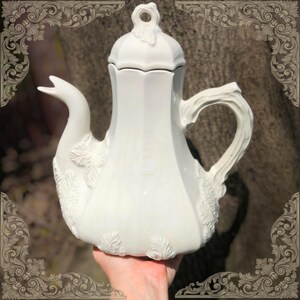May include: A white ceramic teapot with a swan-shaped spout and a branch-like handle. The teapot has a lid with a small loop and is decorated with leaf patterns. The teapot is held in a hand.