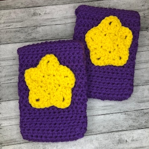 Pode incluir: Duas pulseiras de crochê roxas, cada uma com uma aplicação de estrela amarela brilhante. As pulseiras são feitas de um fio grosso e texturizado, e as estrelas são centralizadas na frente. O fundo é de madeira cinza claro.