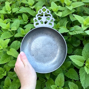 May include: A circular, matte gray metal bowl featuring a decorative, ornate handle with intricate cut-out patterns. The bowl has a smooth, shallow interior surface and a weathered, metallic finish, showcasing a distinct, symmetrical design at the top.