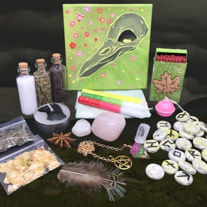 May include: A green canvas with a black crow skull design, pink flowers, and a white feather.  A set of runes, a pink bell, a small box of matches, a white candle, a pink candle, a green candle, a small glass bottle with white salt, a small glass bottle with dried herbs, a small glass bottle with black seeds, a small black candle, a star anise, a clear quartz crystal, a rose quartz crystal, a gold chain with a pentagram pendant, and two small bags of dried herbs.