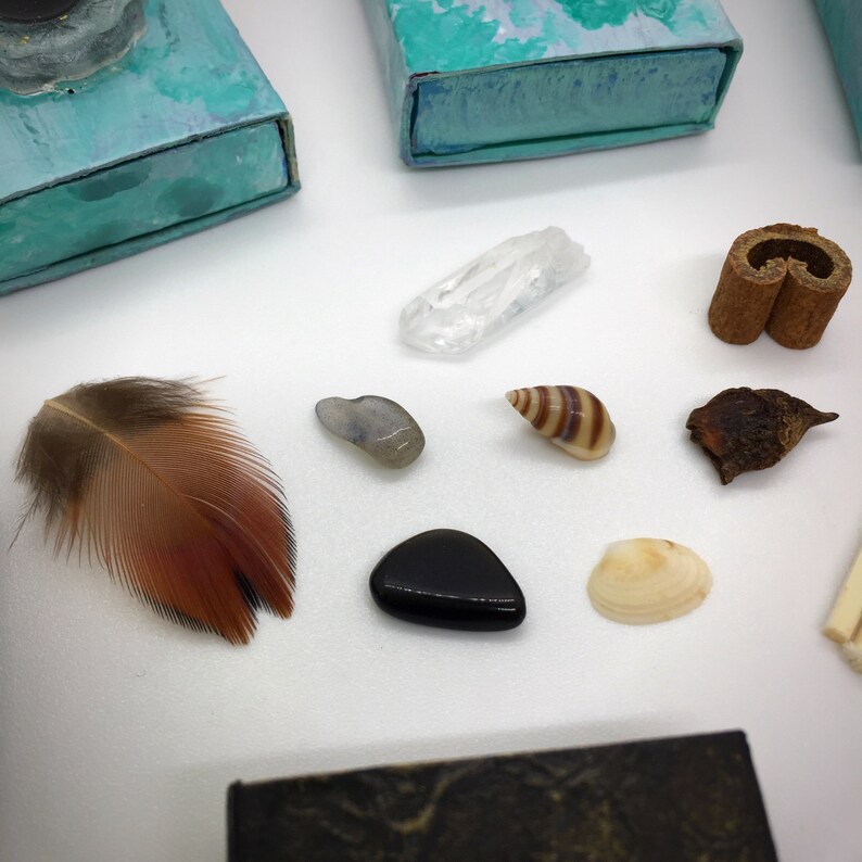 May include: An assortment of natural elements including a brown and red feather, clear quartz crystal, polished black stone, seashells, and cinnamon sticks. Also, two teal and blue painted boxes are visible.