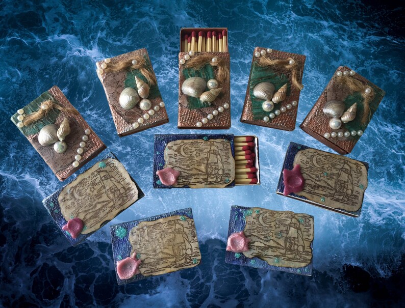 May include: Ten decorative matchboxes with a nautical theme.  The matchboxes are decorated with seashells, pearls, and a nautical map design.  The matchboxes are arranged in a grid pattern on a blue and white background.