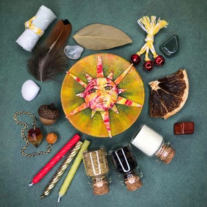 May include: An assortment of spiritual items, including a sun-faced decorative disc, feathers, crystals, candles, and small glass jars with cork stoppers. The items are arranged on a teal surface.