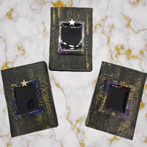 May include: Three handmade, textured black books with gold accents. Each book features a rectangular, black center framed with iridescent and purple resin, topped with a gold star. The books are arranged on a marble-patterned surface.