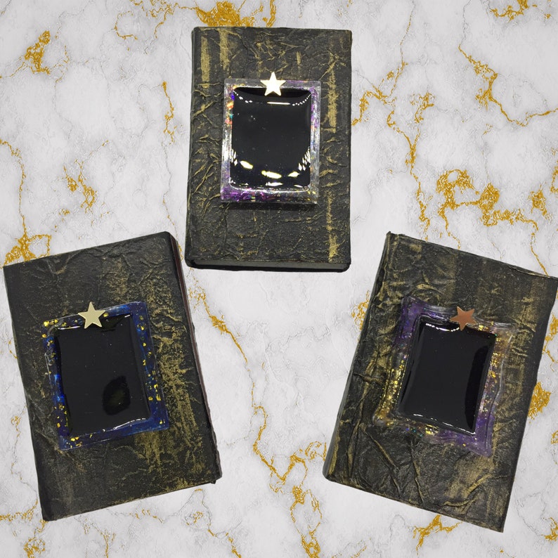 May include: Three handmade, textured black books with gold accents. Each book features a rectangular, black center framed with iridescent and purple resin, topped with a gold star. The books are arranged on a marble-patterned surface.