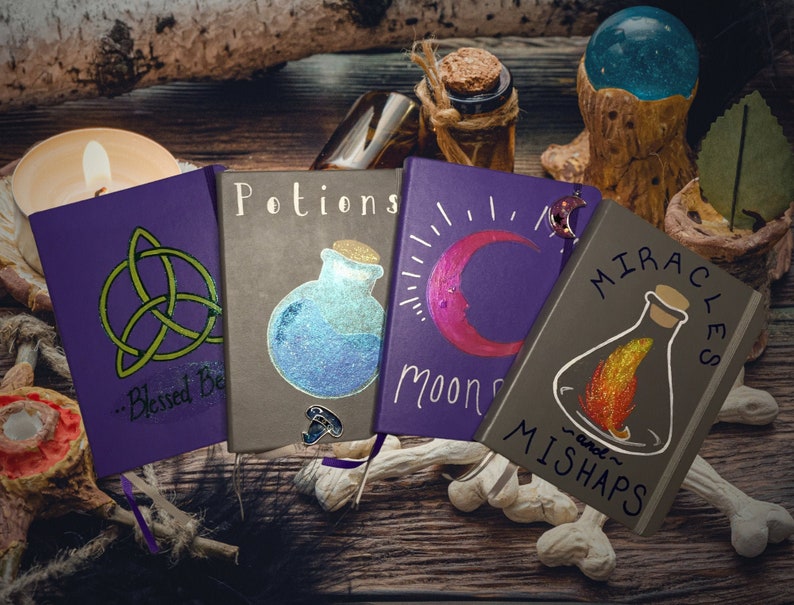 May include: Four purple and gray journals with different designs and titles. The journals are on a wooden surface with a candle, a glass orb, and bones. The titles of the journals are "Blessed Be", "Potions", "Moon Spells", and "Miracles and Mishaps".