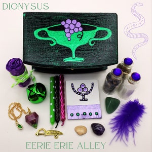 Könnte beinhalten: Eine grüne Holzkiste mit einem gemalten Bild eines Kelches mit Trauben, einer Schlange und dem Text "DIONYSUS". Die Kiste ist von verschiedenen Gegenständen umgeben, darunter eine violette Feder, ein grüner Stein, eine kleine Flasche mit einem violetten Stopfen, eine violette und weiße Kerze, eine grüne Glocke, eine goldene Kette mit einem violetten Kristallanhänger und eine kleine violette und weiße Karte mit einem Symbol darauf. Der Text "EERIE ERIE ALLEY" steht unter der Kiste.