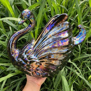 Puede incluir: Una figura de cisne de cerámica con un acabado negro iridiscente brillante que presenta reflejos resplandecientes en azul, violeta y dorado. El cisne se muestra con el cuello curvado con gracia y las alas desplegadas, exhibiendo texturas de plumas moldeadas detalladas.