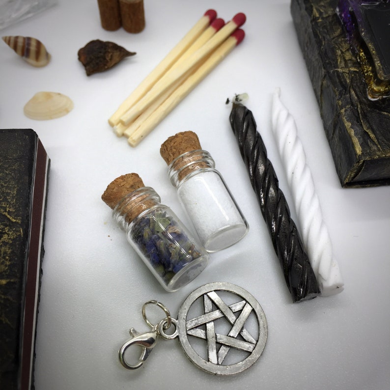 May include: An assortment of spiritual items, including small glass bottles with cork stoppers, filled with herbs and salt. Also, matches, candles, a pentagram charm, and seashells are visible. The items are arranged on a white surface.