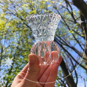May include: A clear, decorative glass perfume bottle with a textured, diamond-like pattern on the top and a rounded base. The bottle is held in a hand against a backdrop of green foliage and a blue sky.