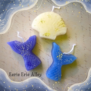 May include: Three decorative candles shaped like a seashell and two mermaid tails. The seashell candle is white with gold glitter. The mermaid tails are blue and purple with gold glitter. The candles are on a light brown background with a blue and white pattern.