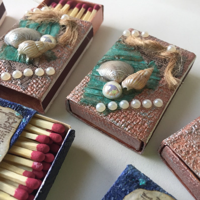 May include: Four small matchboxes decorated with seashells, pearls, and green and brown paper. The matchboxes are arranged in a row on a white surface.