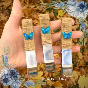 May include: Three silver metal butterfly incense holders wrapped in brown twine with blue glitter butterflies attached. The holders are being held in a hand.