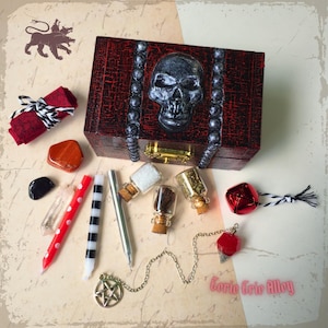 May include: A red wooden box with a silver skull design on the lid. The box is open and contains various items, including small glass bottles, candles, crystals, and a pendulum. The text "Corie Erie Alloy" is visible on the bottom right corner of the image.