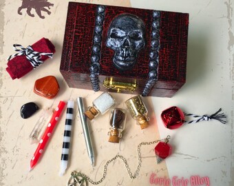 May include: A red wooden box with a silver skull design on the lid. The box is open and contains various items, including small glass bottles, candles, crystals, and a pendulum. The text "Corie Erie Alloy" is visible on the bottom right corner of the image.