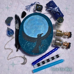 Pode incluir: Um círculo de madeira pintado de azul e preto com uma silhueta de corvo voando na frente de uma lua cheia. O círculo é cercado por vários itens, incluindo um sino de prata, um rolo de tecido azul e branco, uma corrente de prata com um amuleto de lua crescente, uma pena preta, um cristal transparente, um pêndulo de cristal azul, duas pequenas garrafas de vidro com ervas secas e duas velas azuis.