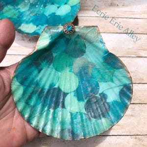 May include: Handmade seashell with a teal and turquoise design. The shell is decorated with a pattern of overlapping circles in shades of blue and green. A small, round, blue gem is attached to the top. The words "Eerie Erie Alley" are in the upper right corner.