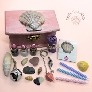 May include: A pink wooden box with a seashell on the lid, containing a variety of items including a small rolled cloth, three small bottles, a necklace with a mermaid pendant, a pink crystal, a seashell, a bell, and three candles. The box is labelled "Eerie Erie Alley".
