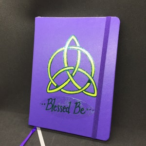 May include: Purple notebook with a green and yellow triquetra symbol and the text "...Blessed Be..." on the front cover.