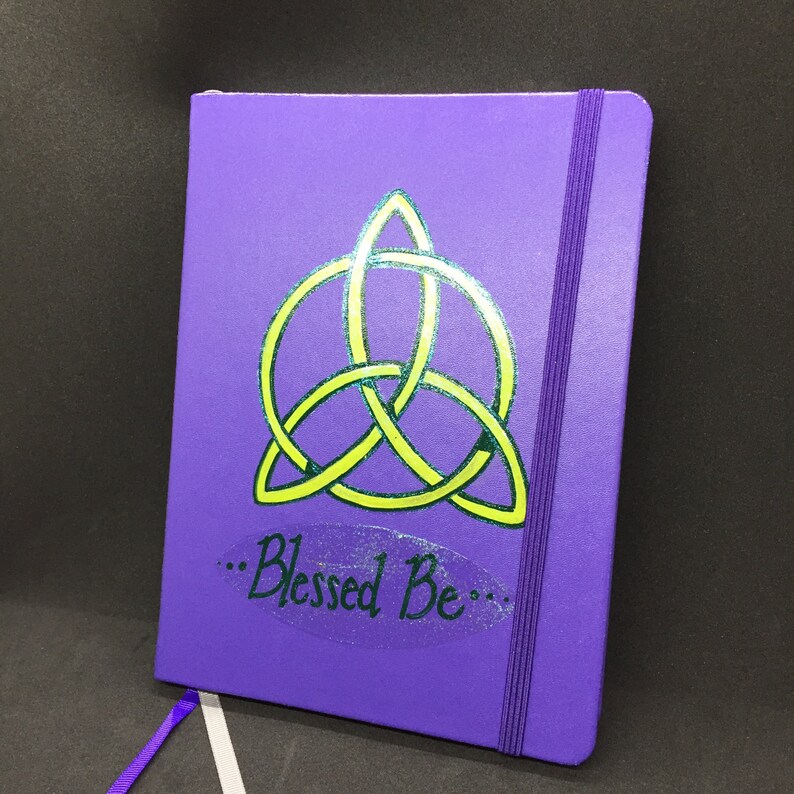 May include: Purple notebook with a green and yellow triquetra symbol and the text "...Blessed Be..." on the front cover.