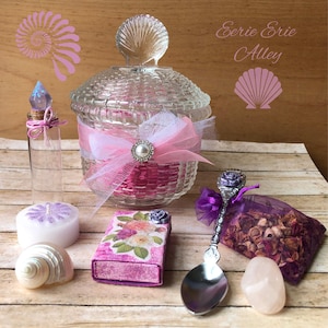 Puede incluir: Una colección de artículos decorativos, que incluyen un tarro de cristal con tapa en forma de concha, una botella pequeña con tapón de cristal, una vela, una concha, una caja de cerillas, una cuchara, una bolsa de popurrí y una piedra de cuarzo rosa. Los artículos son en tonos rosa, morado y transparente.