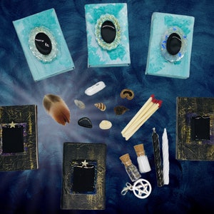 May include: An assortment of mystical items arranged on a dark blue background. The items include three teal-colored books with oval embellishments, three black books with star accents, a feather, stones, shells, matches, candles, and small bottles.