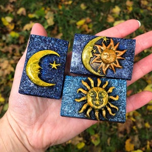 Könnte beinhalten: Drei rechteckige Kunstwerke zum Thema Himmelskörper. Jedes zeigt ein goldfarbenes Sonnen-, Mond- und Sterndesign auf einem strukturierten, dunkelblauen Hintergrund. Sonne und Mond haben Gesichter. Die Stücke werden in einer Hand gehalten.