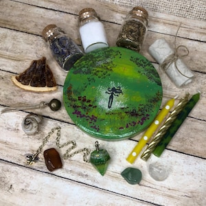 May include: An assortment of spiritual items, including small glass jars with cork stoppers, a green painted wooden disc with a figure, candles, crystals, and a pendulum. The items are arranged on a light wood surface.