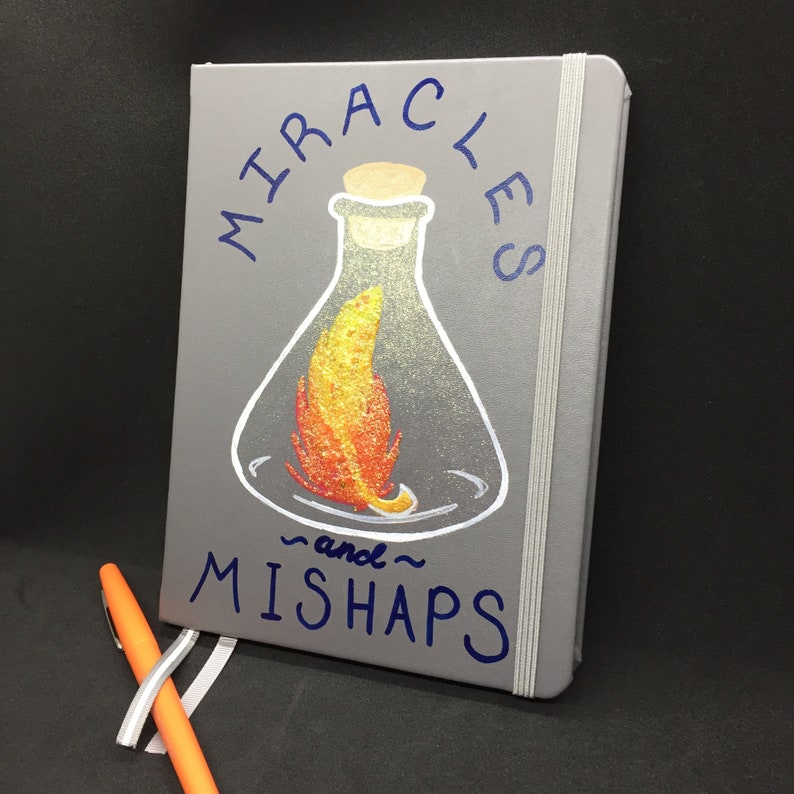 May include: Gray notebook with a blue inscription that reads "Miracles and Mishaps". The notebook has a white elastic band closure and a drawing of a glass flask with a flame inside.